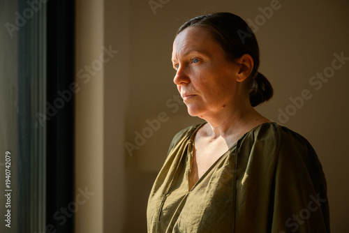 Fotografie Thoughtful middle-aged woman standing by a window in warm natural light, looking outside with a serious contemplative expression, quiet interior portrait
