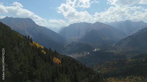Golden trees blanket mountain slopes. Distant peaks rise under scattered clouds. Vibrant valley glows in late autumn light. No people, only nature's quiet majesty