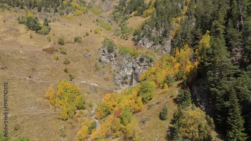 Autumn mountainside reveals golden, green, and brown hues. Rocky cliffs frame a winding trail through colorful trees. Aerial view shows nature's vibrant seasonal transformation
