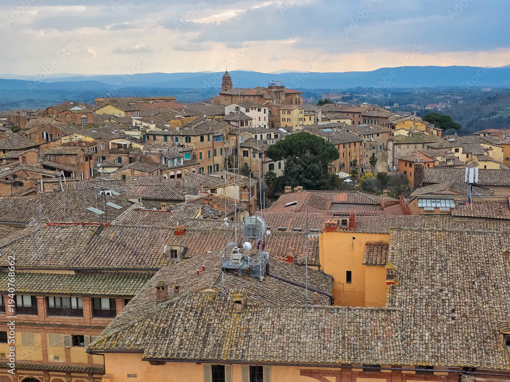 Fototapeta premium Aerial View of Medieval Rooftops in Siena, Italy. Scenic Cityscape of Tuscan Town with Terracotta Tiles and Historic Architecture. Panoramic Skyline of Siena's Ancient District in Tuscany