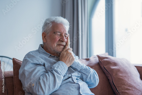 Obraz na plátně An older man sits alone on a sofa pressing his cheek due to a severe toothache,