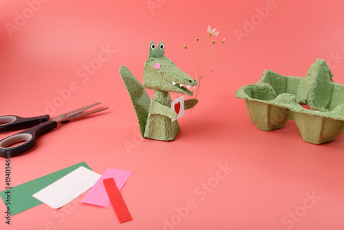 Handmade alligator character from recycled egg box holding heart card and flower for eco brand marketing green business promotion sustainability workshop social media content creative craft concept