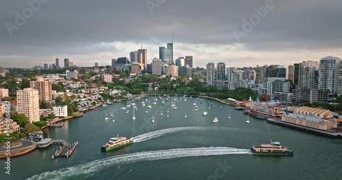 Sydney cityscape and North Sydney suburbs flanking Lavender Bay, calm inlet filled with moored yachts, two ferries navigate the water, creating wakes under a cloudy sky. Aerial drone zoom out panorama