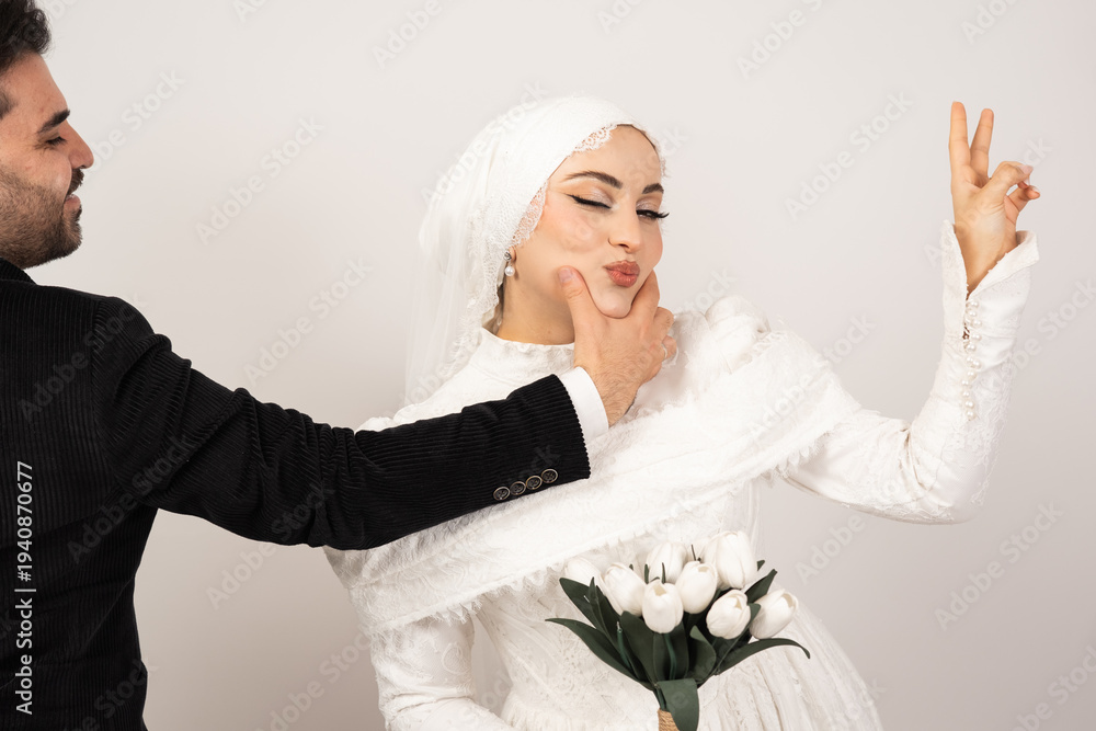 Naklejka premium Playful wedding portrait of a cheerful bride and groom captured in a studio with a minimal background. The groom gently holds the bride’s chin while she makes a funny kissing face 
