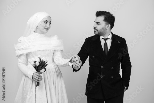 Black and white studio portrait of a bride and groom sharing a romantic moment on their wedding day. The elegant bride in a modest white wedding dress and veil gently holds the groom’s hand 