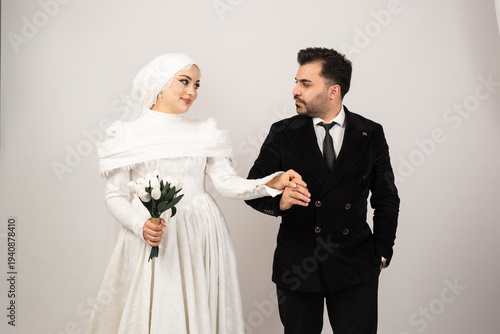 Elegant studio portrait of a Muslim bride and groom holding hands and looking at each other with love. The bride wears a modest white lace wedding dress with hijab and holds a bouquet of white tulips