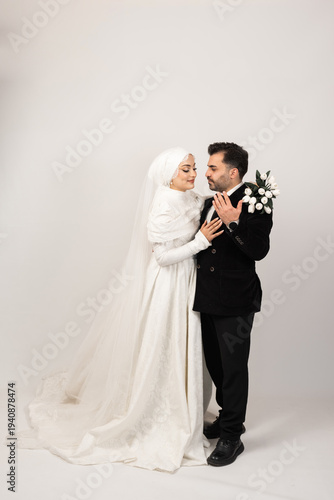 Romantic studio portrait of a Muslim bride and groom standing close together. The bride in a modest white lace wedding dress and hijab gently touches the groom while he holds a bouquet of white tulips