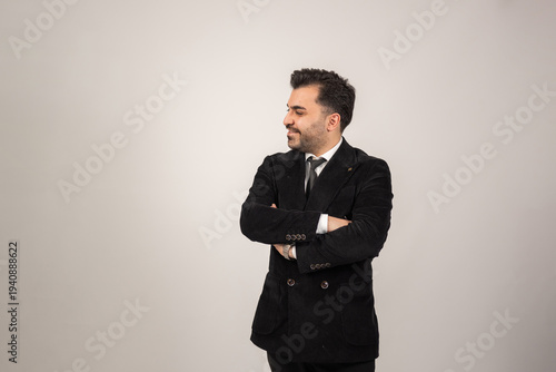 Studio portrait of a confident groom standing with crossed arms in a classic black suit and tie. Minimal background emphasizes elegance, style, and modern masculinity.