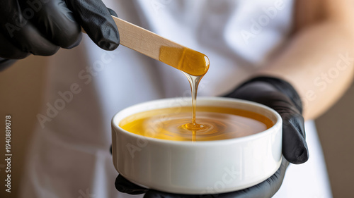 A person in black gloves holding a bowl of golden wax with a wooden spatula dripping more wax, ready for hair removal.