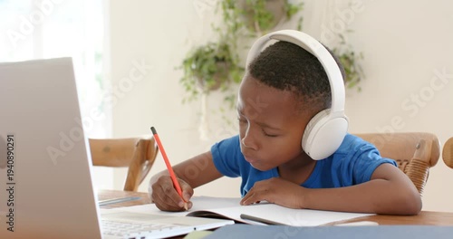 African American child writing notebook with pencil at home with headset raising hand to laptop cue