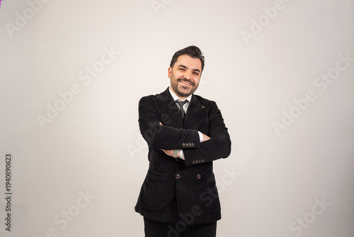 Confident young man standing in a minimalist studio with arms crossed and smiling at the camera. He is wearing a stylish black suit with a white shirt and tie, creating a modern and elegant look