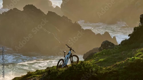Mountain bike resting on a grassy cliffside. Overlooking the dramatic coastline of Portugal with golden sunlight illuminating the powerful ocean waves crashing against rocky formations during a sunset