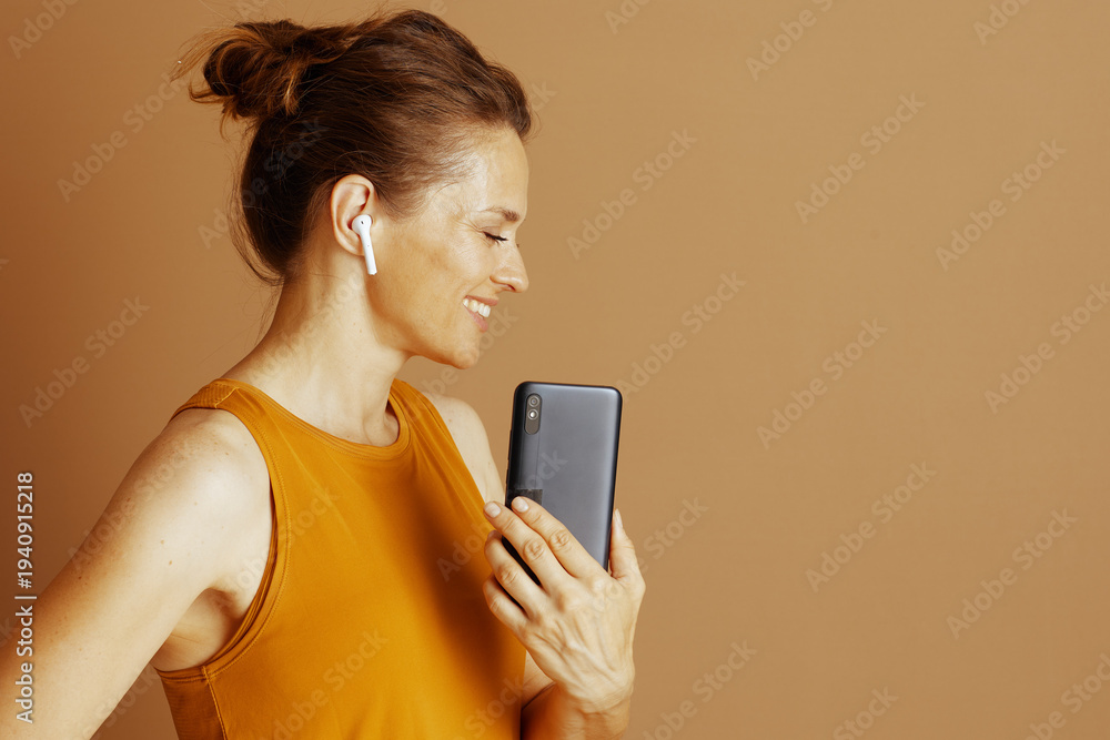 Fototapeta premium A woman with bun, in mustard yellow top, smiles serenely with closed eyes, listening through earbuds and holding smartphone. It captures moment of modern relaxation and personal digital connection
