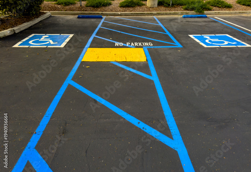 Accessible parking spaces with blue lines and symbols flank a striped no parking zone with a yellow ramp on a clean asphalt lot bordered by greenery and curbs