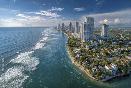 Aerial cityscape of modern coastal town with high rise apartment buildings along shoreline under partly cloudy blue sky, showing waves and urban greenery in vibrant atmosphere