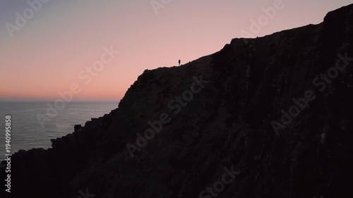Silhouette of man standing on high coastal cliff in Costa Vicentina Natural Park near São Teotónio Odemira Alentejo Portugal as drone pushes forward revealing Atlantic Ocean horizon at sunset.