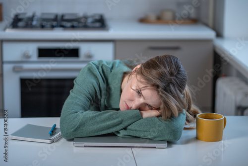 Photos Distressed exhausted young woman feeling tired, frustration while working or studying from home lying head on laptop at kitchen table looking with tension