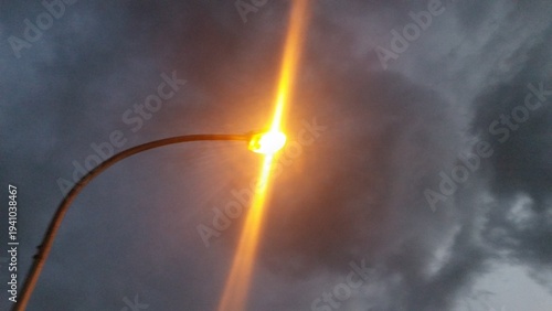 Foto An orange-yellow streetlight glowing against dark cloudy skies, its light forming vertical beams upward and downward due to atmospheric diffraction during dusk