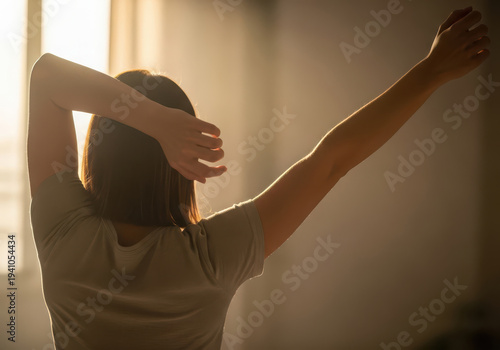 Woman stretching arms up in morning light near window.