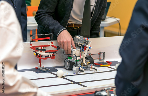 A student in a school uniform is fine-tuning a custom-built robot during a technical workshop or competition. The robot is constructed from modular building blocks, gears, motors, and electronic senso