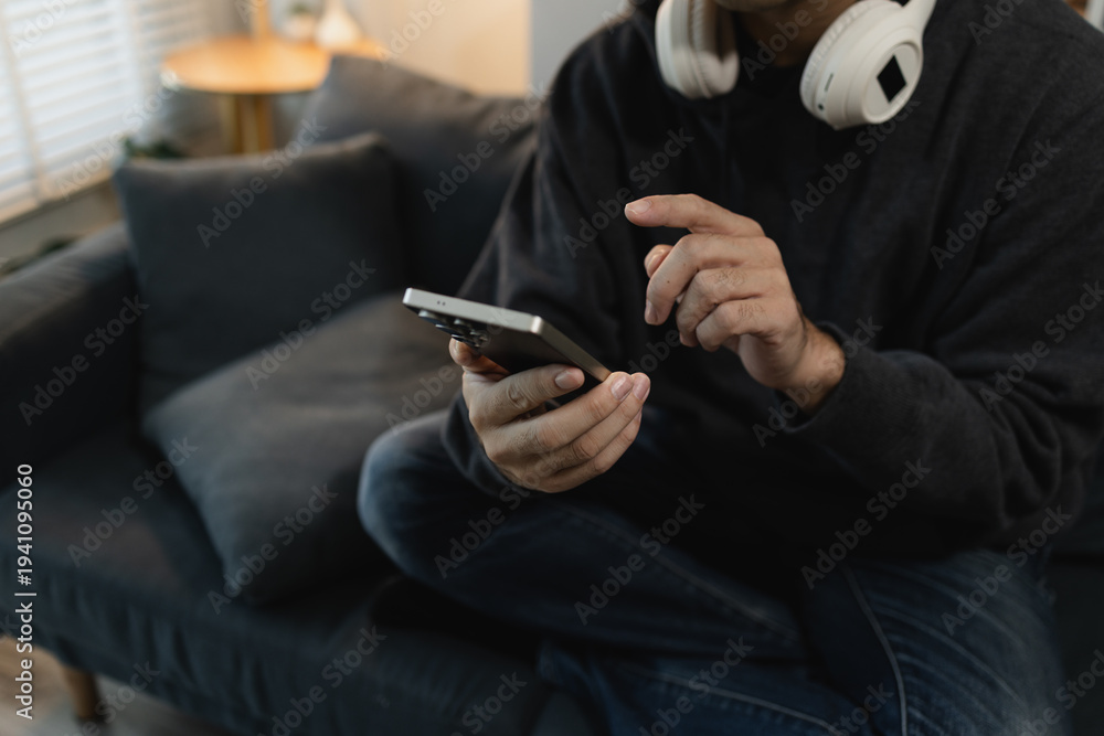 Fototapeta premium Man using smartphone while relaxing on sofa at home, browsing mobile apps and digital content, representing modern lifestyle, online communication and mobile technology.