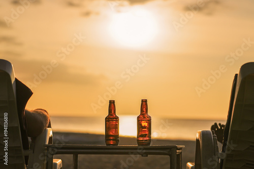 Konstfotografi Two bottles of beer are on the table at sunset on the beach - Two friends or lovers are watching the sunset sitting in chairs back view