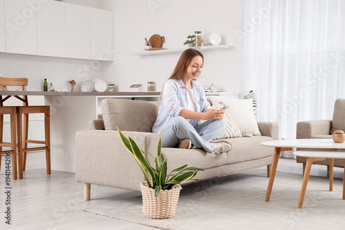 Beautiful young woman sitting on sofa and using mobile phone in living room