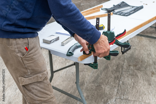 Photos Man in casual clothes clamps a wooden plank to a folding worktable, focused on p