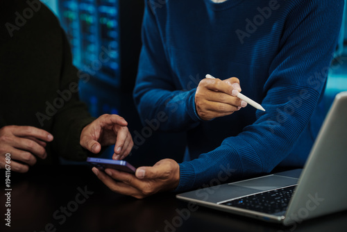 Close-up of two coworkers collaborating, using smartphone and stylus beside laptop in modern office, blue lighting, tech and productivity concept