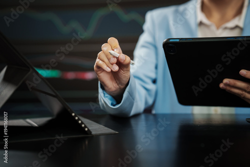 Business professional holding tablet and stylus at desk with laptop, blurred stock market chart background, modern finance and technology concept.