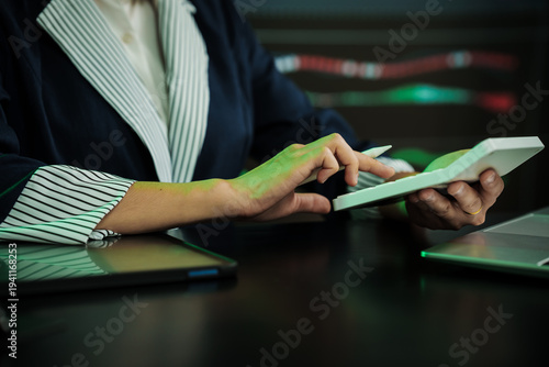Close-up of businesswoman in striped blazer using tablet at desk, with laptop and green ambient light, modern office setting, technology and finance concept.