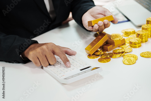 Businessperson calculating wealth with calculator, holding gold bar; stacks of gold bars and coins on office desk near laptop, finance investment concept.