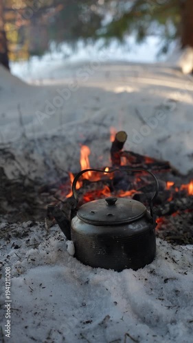 Metal camping kettle heating on a campfire in a snowy winter forest. Bushcraft outdoor cooking and wilderness survival concept with warm flames, cold snow