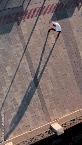 Young athlete wearing a red beanie and white sweatshirt stretching his arm and shoulder before a run near a city bridge. The urban setting and early daylight highlight preparation for an outdoor