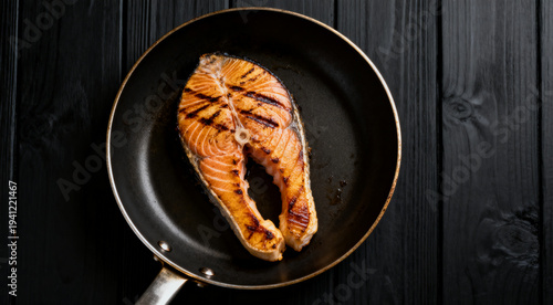 Frying pan with grilled salmon steak over black wooden surface