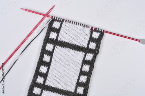A hand knitted piece of fabric features a creative design mimicking a black and white vintage movie film strip on red knitting needles