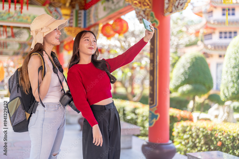Obraz premium Two happy asian women tourists taking selfie with smartphone at Chinese temple, Young backpacker friends waving to video call on summer vacation holiday, Culture travel and technology concept