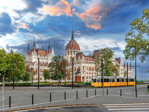 A yellow historic tram passes in front of the ornate Hungarian Parliament Building under a dramatic sunset sky in Budapest.