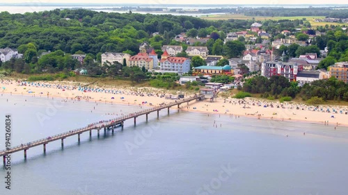 An aerial view shows the Binz pier on Rügen stretching into the Baltic Sea, lined with a busy sandy beach and classic resort architecture.
