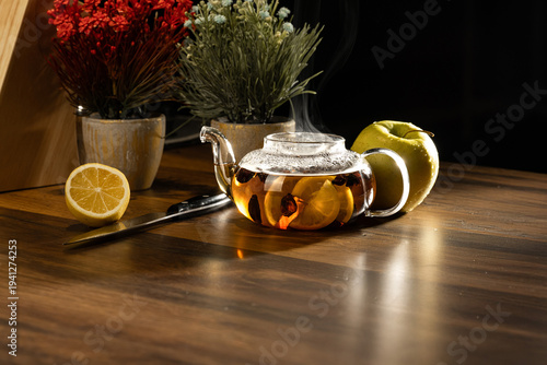Cozy home tea ritual with steaming glass teapot, fresh lemon, apple, and decorative flowers on a wooden table