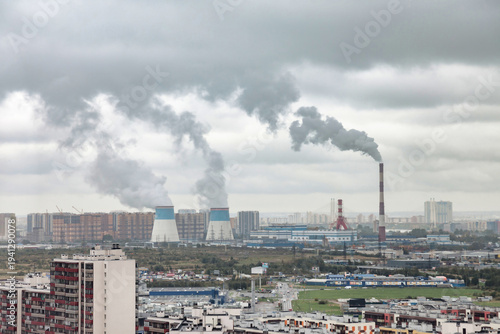 Tall chimney emitting smoke in the distance. Cooling towers and buildings present. The sky is cloudy over a cityscape. There is copy space in the sky.