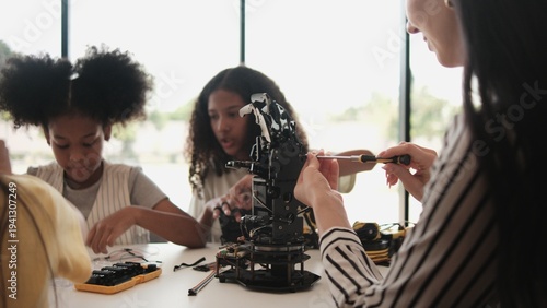 Group of student girls robotic arms design and experiment with female teacher instructing at learning table in primary science classroom, discussing technology knowledge for kids of elementary school.