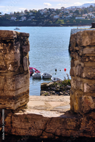 View of weathered stone embrasure framing boats bobbing gently on the tranquil bay, contrasting with the distant hillside homes, Saint-Jean-de-Luz, Nouvelle-Aquitaine, France.