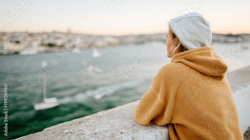 A thoughtful woman in a cozy hoodie observes the stunning waterfront view at sunset, reflecting on life’s moments, embodying serenity and connection with nature.
