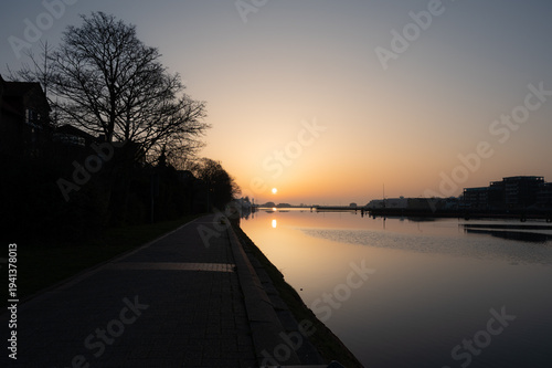 A tranquil harbor scene at sunrise with a footpath and trees silhouetted along the shore; the sunrise view from the dike bridge in 26382 Wilhelmshaven, Germany.