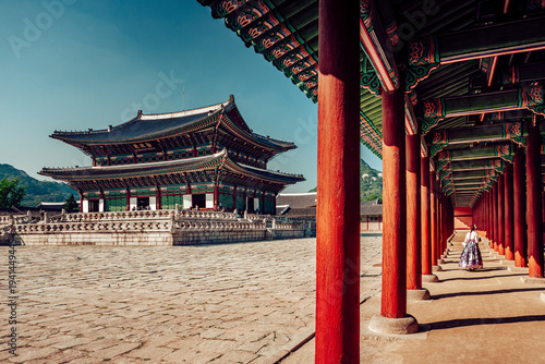 View of a woman in traditional dress walks through a corridor with vibrant red pillars, contrasting with the grand palace and expansive courtyard, Seoul, Seoul, South Korea.