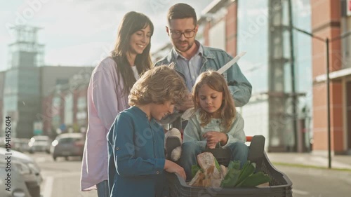 Family with two kids checking shopping receipt outside. Man holding bill, woman standing nearby, children looking at groceries in cart with bread and vegetables in parking lot after supermarket visit.