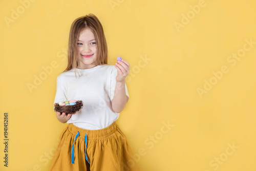 Little girl with long brown hair holds colorful Easter eggs in a nest while smiling against a bright yellow background, wearing a white shirt and yellow skirt
