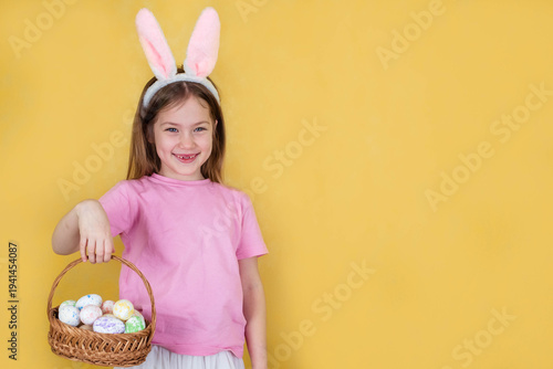 Little girl wearing pink bunny ears holds a wicker basket filled with colorful Easter eggs against a bright yellow background, smiling cheerfully
