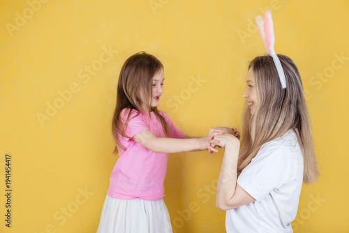 Caucasian woman and girl wearing bunny ears interact playfully against a bright yellow background, showcasing a festive and cheerful atmosphere for Easter celebrations